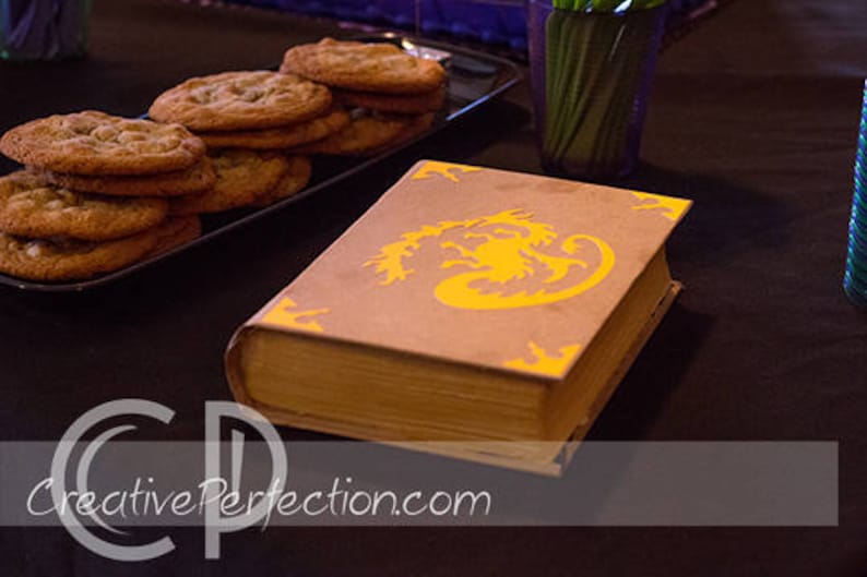May include: A brown book with a yellow dragon design on the cover. The book is sitting on a black table with a plate of cookies in the background.