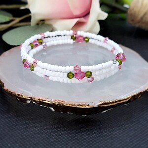 May include: A white beaded bracelet with pink and green crystal accents. The bracelet is wrapped around itself twice and is sitting on a white stone surface.