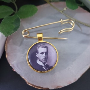 May include: A gold-coloured safety pin with a circular pendant featuring a black and white portrait of a man. The pendant has a gold-coloured border. The pin is resting on a light-coloured, round surface.