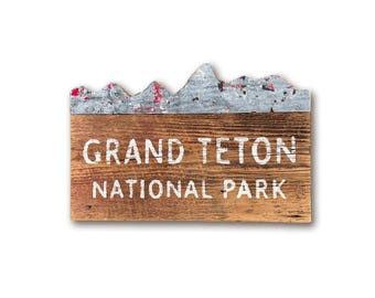 May include: A rustic wooden sign with a metal mountain range design at the top. The sign reads "Grand Teton National Park" in white paint.