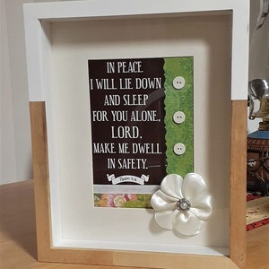 May include: A white frame with a brown wood border, containing a framed print with a green background and white buttons. The print features a religious quote: "In peace I will lie down and sleep for you alone, Lord, make me dwell in safety." The quote is attributed to Psalm 4:8. A white satin flower with a pearl button in the center is attached to the bottom right corner of the frame.