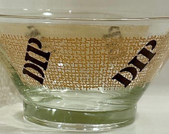 Puede incluir: Un bol de vidrio transparente para dip con un diseño estampado en dorado y marrón. Las palabras "Dip" están impresas en ambos lados del bol en negro.