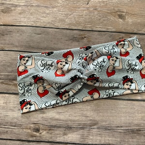 May include: A white headband with a black polka dot pattern and illustrations of a woman flexing her arm with the words "Scrub Life" in a heart-shaped bubble. The woman has red hair and a red top.
