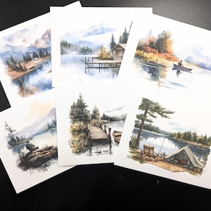 May include: A collection of watercolor landscape art prints featuring serene lake scenes with mountains, trees, and cabins. Each print showcases a different view, including a fisherman, a dock, and a tent, all in soft blues, greens, and browns.