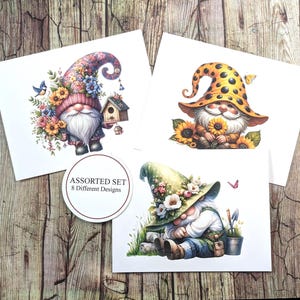May include: An assortment of three square cards featuring whimsical gnome illustrations. Each gnome is depicted with a long white beard and a colorful hat, surrounded by flowers and butterflies. The cards are on a wooden background.