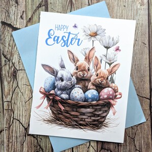 May include: Happy Easter greeting card featuring three brown and white bunnies in a wicker basket with colorful Easter eggs. The bunnies are surrounded by white daisies and butterflies.