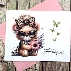 May include: A greeting card featuring a cartoon bear wearing sunglasses and a leopard print scarf, holding a bouquet of flowers. The card has the text "Thinking of you" and several green butterflies.