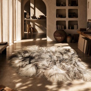 May include: A large, long-haired sheepskin rug featuring a natural color palette of cream, beige, and dark charcoal gray. The rug has a thick, shaggy texture with irregular edges, showcasing the organic, flowing shape of the animal hide.