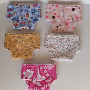 Puede incluir: Cinco pañales para muñecas con diferentes estampados. Los pañales están hechos de tela y tienen un ribete blanco. Los estampados incluyen cerezas y fresas sobre un vichy azul y blanco, lunares marrones y rosas, amarillo con pequeños lunares morados y blancos, blanco con pequeñas flores rosas y azules, y rosa con flores de hibisco blancas.