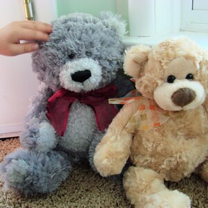 May include: Two plush teddy bears, one gray and one beige, are sitting next to each other. The gray bear has a red ribbon around its neck. The beige bear has a yellow and orange ribbon around its neck.