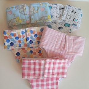 May include: A collection of colorful cloth diapers. Designs include polka dots, hearts, and gingham patterns. The diapers have adjustable closures and are made of soft fabric. These reusable diapers are a sustainable alternative to disposable options.