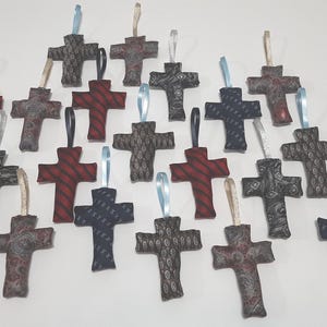 May include: A collection of decorative cross ornaments in various colors and patterns. The crosses are made from fabric and have ribbons for hanging. The colors include red, black, and gray.