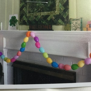 Puede incluir: Una guirnalda colorida de huevos de Pascua de plástico cuelga sobre una repisa de chimenea blanca. La guirnalda está hecha de huevos de colores pastel, incluyendo rosa, morado, azul, verde, amarillo y naranja. La guirnalda está enhebrada con un hilo transparente. La repisa de la chimenea es blanca y tiene un marco de ladrillo. La guirnalda es una decoración festiva para Pascua.