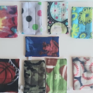 May include: A selection of rectangular fleece fabric items with diverse patterns. Designs include tie-dye, polka dots, floral, basketball, camouflage, football, and baseball. Each item has a small white tag attached.