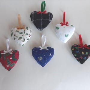 May include: A collection of heart-shaped Christmas ornaments. The ornaments feature various patterns, including holly, plaid, and a starry night design. Each ornament has a ribbon for hanging and a small bow.