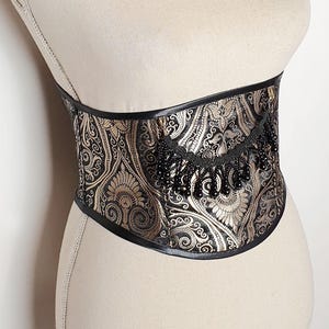 Op de afbeelding: Een goudkleurig en zwart korset met een sierlijk paisley patroon. Het korset wordt op een mannequin getoond. De boven- en onderranden zijn afgewerkt met zwart materiaal. De tekst "My Oppa" is zichtbaar.