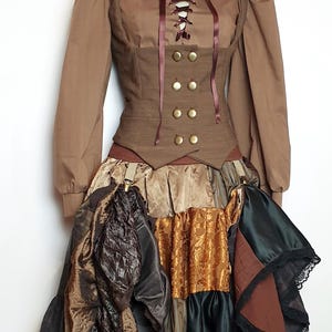 Peut inclure: Une tenue de style steampunk marron sur un mannequin. L'ensemble comprend un haut à col avec un corsage lacé, des boutons dorés et une jupe superposée avec diverses textures et couleurs, dont le noir, l'or et le marron. Fait main.