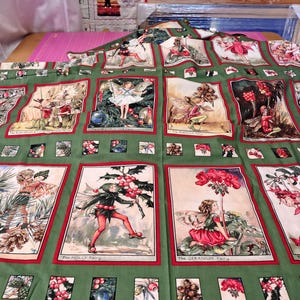 May include: A green fabric panel featuring a grid of rectangular illustrations of various fairies, including the Pine Tree Fairy, Holly Fairy, Geranium Fairy, and Fuchsia Fairy, each labelled with text at the bottom of their respective frames.