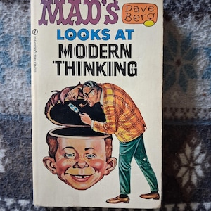May include: Vintage paperback book titled "MAD's Looks at Modern Thinking" by Dave Berg. The cover features a cartoon illustration of a man with a magnifying glass examining the open head of another cartoon character. The book has a cream-colored background with purple and black text.