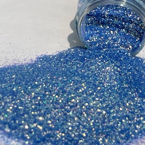 May include: A close-up of a jar of light blue glitter with iridescent shimmer. The glitter is spilling out of the jar onto a white surface.