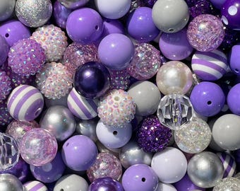 May include: A collection of purple beads in various shades and textures. The beads include solid, glittered, striped, and crystal-like designs. Colors range from light lavender to deep violet, with silver and white accents.