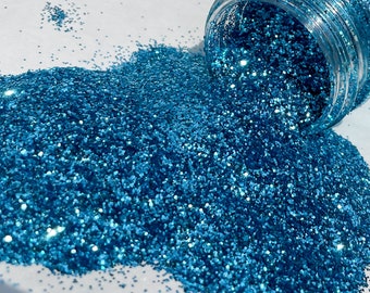 Op de afbeelding: Een close-up van een potje met teal blauwe glitter, met wat glitter dat uitloopt op een wit oppervlak.