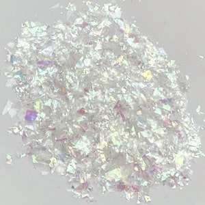 May include: A pile of iridescent white glitter flakes. The glitter is made of small, irregular pieces of plastic that reflect light in a rainbow of colors.