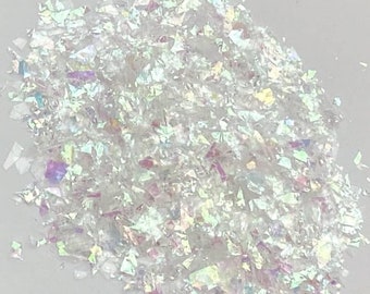 May include: A pile of iridescent white glitter flakes. The glitter is made of small, irregular pieces of plastic that reflect light in a rainbow of colors.