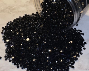 以下が含まれることがあります： 透明なプラスチック容器から、たくさんの黒いラインストーンがこぼれ出ています。ラインストーンは小さく丸く、光沢のある仕上げになっています。