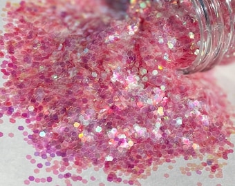 Op de afbeelding: Een close-up van roze, iriserende glitter, die uit een doorzichtige pot op een wit oppervlak stroomt.