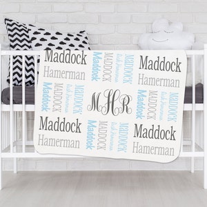 May include: White baby blanket with the names Maddock and Hamerman repeated in a blue and gray pattern. The blanket also has the monogram MHR in gray.