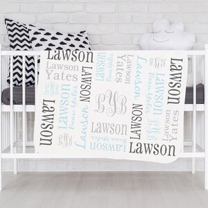 Op de afbeelding: Witte babydeken met de namen Lawson en Yates die in een blauw en grijs lettertype worden herhaald. De deken heeft ook de initialen "BYB" in een grijs lettertype.