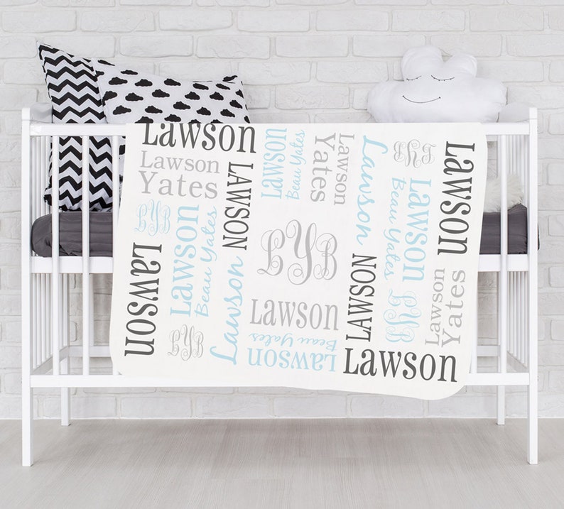 Op de afbeelding: Witte babydeken met de namen Lawson en Yates die in een blauw en grijs lettertype worden herhaald. De deken heeft ook de initialen "BYB" in een grijs lettertype.