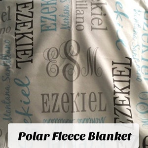 Op de afbeelding: Een wit fleece deken met de naam "Ezekiel" bedrukt in grijze en blauwe letters. Het deken heeft ook een monogram met de letters "E" en "M" in grijs.