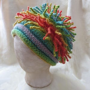 May include: A colorful, hand-knitted hat with a unique design. The hat features a rainbow of colors, including green, blue, yellow, orange, and red. The top of the hat has a textured, spiky design. The hat is displayed on a mannequin head.