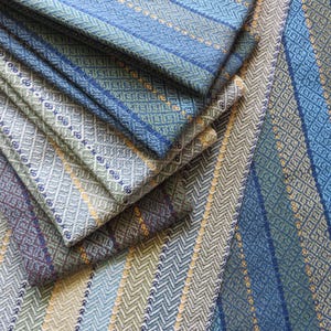 May include: Close-up of folded fabric samples in shades of blue, green, and gray. The textiles feature intricate geometric patterns and stripes, with a mix of textures and designs. The fabrics are likely for home decor or fashion.