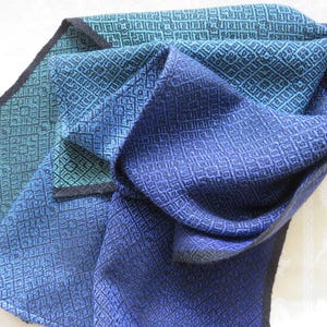 以下が含まれることがあります： 幾何学模様の青とターコイズ色の織りスカーフ。 スカーフは折りたたまれており、生地のさまざまな色と質感が示されています。 エッジは暗いボーダーで仕上げられています。