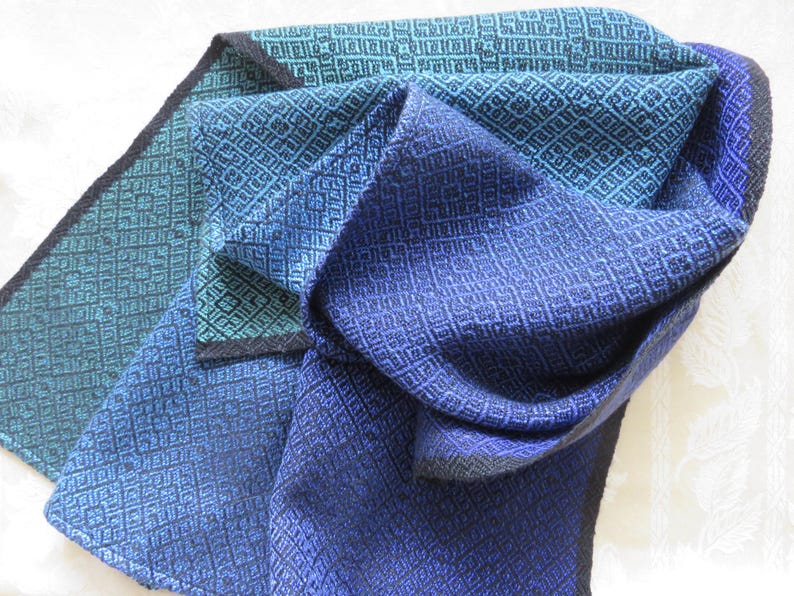 以下が含まれることがあります： 幾何学模様の青とターコイズ色の織りスカーフ。 スカーフは折りたたまれており、生地のさまざまな色と質感が示されています。 エッジは暗いボーダーで仕上げられています。