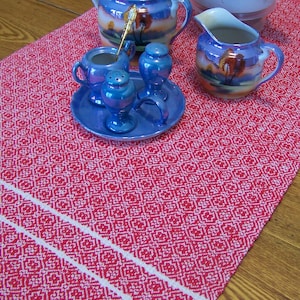 May include: A red and white table runner with a geometric pattern. A blue and white teapot, creamer, and sugar bowl are on the runner. There are also four blue and white salt and pepper shakers.