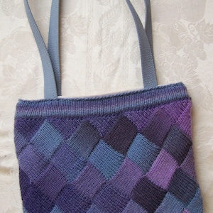 May include: A knitted tote bag with a woven pattern in shades of purple, blue, and gray. The bag has two gray straps.