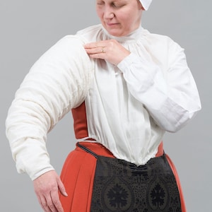 Puede incluir: Una camisa de lino blanca con una manga fruncida y una gorra blanca. La camisa está metida en una falda roja con un delantal estampado negro.