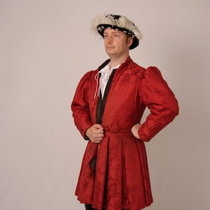 Può includere: Un uomo indossa un cappotto rosso con motivi, una cintura nera e un cappello nero con piume bianche. Indossa calze nere e scarpe bordeaux.