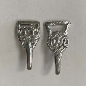 May include: Two silver-toned metal buckles with intricate floral designs. The buckles have a hook-like shape at the bottom.