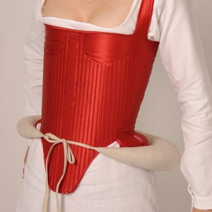 Könnte beinhalten: Ein rotes Satin-Korsett mit einem weißen Baumwoll-Unterhemd und einem weißen Stoffgürtel. Das Korsett hat einen quadratischen Ausschnitt und verstellbare Träger.
