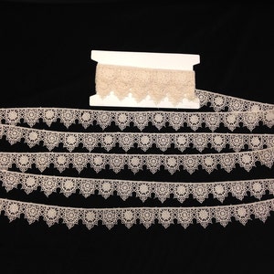 Puede incluir: Una bobina de encaje de color marfil con un borde festoneado y un delicado patrón floral. El encaje está dispuesto en varias filas, mostrando su diseño intrincado.