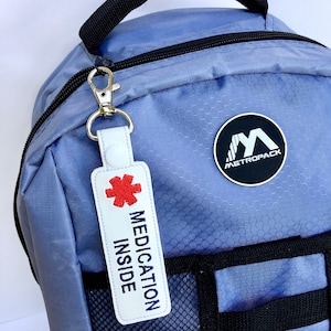 May include: A light blue backpack with a black handle and a white tag that says "MEDICATION INSIDE" with a red medical symbol. The tag is attached with a silver clasp. A black and white "METROPACK" logo is on the backpack.