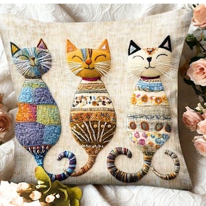 Op de afbeelding: Decoratief vierkant kussen met drie geborduurde kattenontwerpen. De katten hebben patchwork lichamen in blauw, oranje en crème, met geborduurde details en gekrulde staarten. Het kussen staat op een crèmekleurige achtergrond.