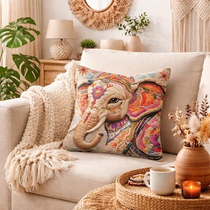 May include: A decorative square pillow with an embroidered elephant design. The elephant is adorned with colorful patterns in orange, pink, and purple. The pillow is on a beige sofa with a cream-colored throw blanket. A coffee table holds a tray and a mug.