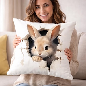 Może przedstawiać: Biała kwadratowa poduszka z jasnobrązowym królikiem wychodzącym z dziury. Królik ma długie uszy i ciemne oczy. Poduszka jest trzymana przez osobę.