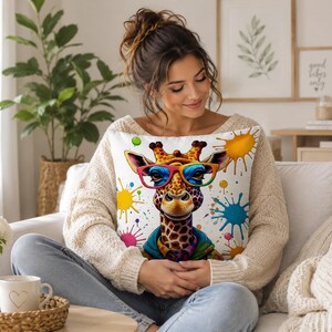 May include: A white throw pillow featuring a cartoon giraffe wearing colorful glasses and a blue jacket. The pillow has colorful paint splatters and is held by a person wearing a cream-colored sweater and blue jeans.